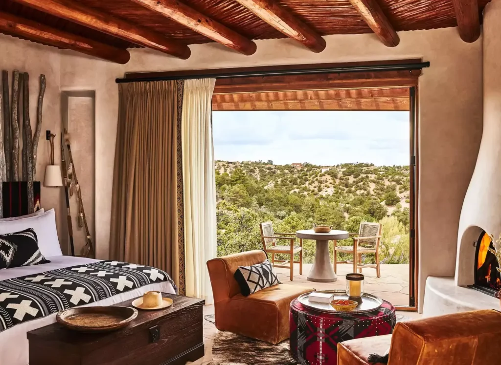 Bishop’s Lodge Bedroom interior featuring a plush bed, cozy sitting area, and panoramic views of lush mountain greenery through a scenic window.