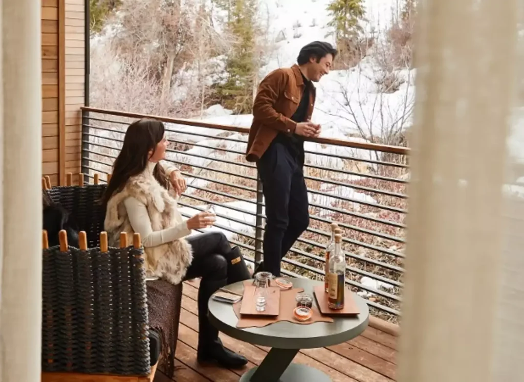 A woman and a man, smiling, with a table of delicacies next to them, while they enjoy the view from a deck of a snow-covered mountain view