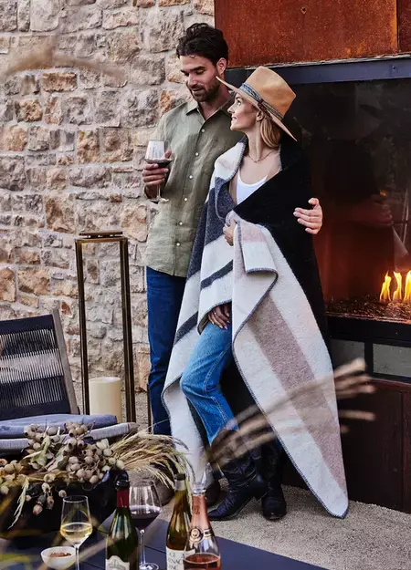 A man embraces over the shoulder a women covered in warm blanket, while he enjoys a glass wine, with a fireplace in the background
