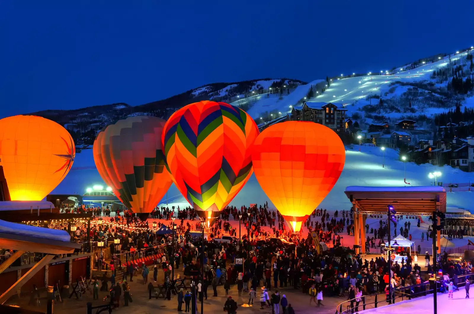Steamboat Springs Night Festival at the base of a snow-covered mountain, with night lights going above the mountain, with the public surrounding four hot air balloons of different colors inside the festival area